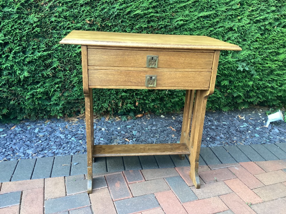 An antique light oak Jugendstil side table featuring two drawers with rectangular brass drop handles, flared side supports, and a lower shelf.