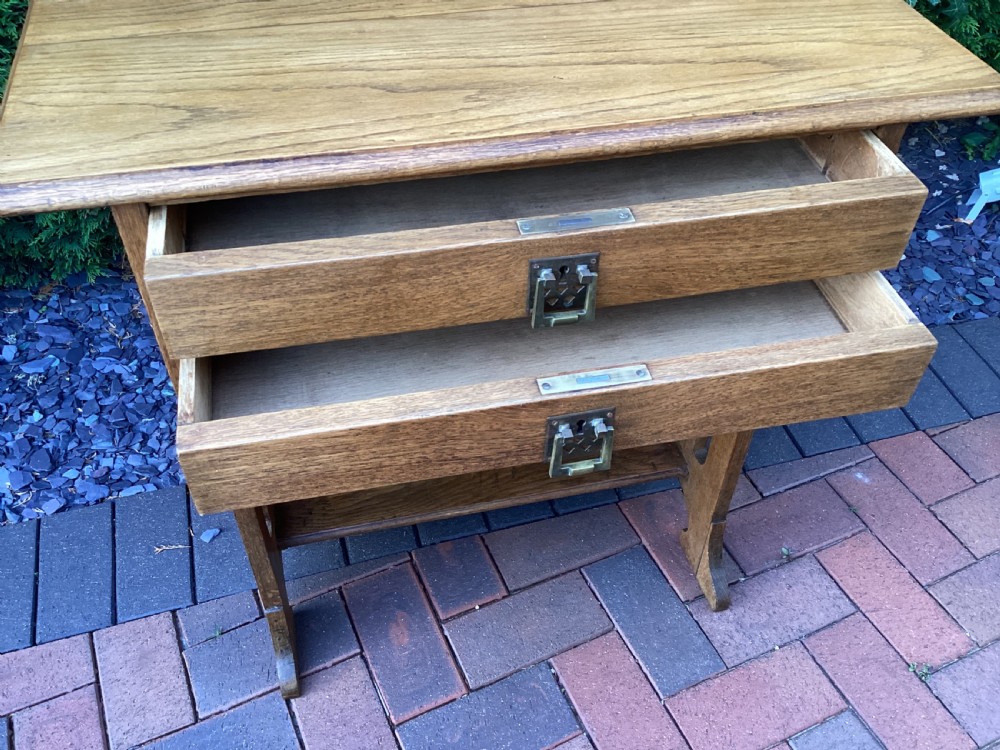 An antique light oak Jugendstil side table featuring two drawers with rectangular brass drop handles, flared side supports, and a lower shelf. View X