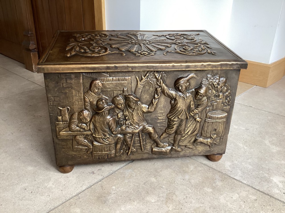 An antique brass-clad wooden fireside log box featuring embossed relief scenes of figures in a tavern on the front and foliate scrollwork on the lid.