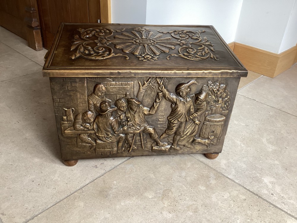 An antique brass-clad wooden fireside log box featuring embossed relief scenes of figures in a tavern on the front and foliate scrollwork on the lid. View II