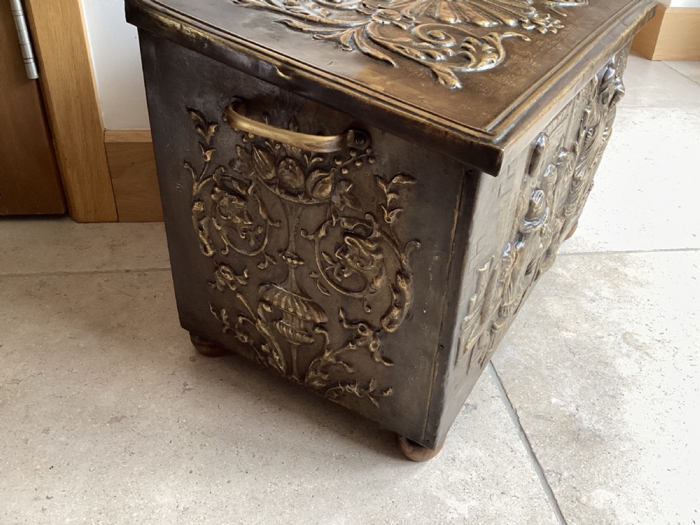 An antique brass-clad wooden fireside log box featuring embossed relief scenes of figures in a tavern on the front and foliate scrollwork on the lid. View III