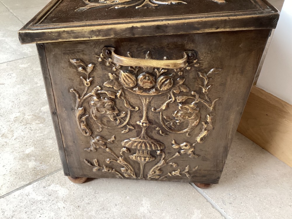 An antique brass-clad wooden fireside log box featuring embossed relief scenes of figures in a tavern on the front and foliate scrollwork on the lid. View V