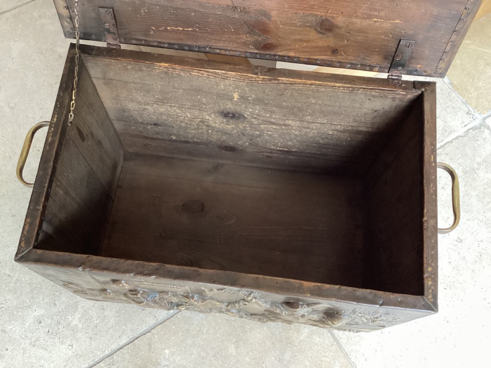 An antique brass-clad wooden fireside log box featuring embossed relief scenes of figures in a tavern on the front and foliate scrollwork on the lid. View VI