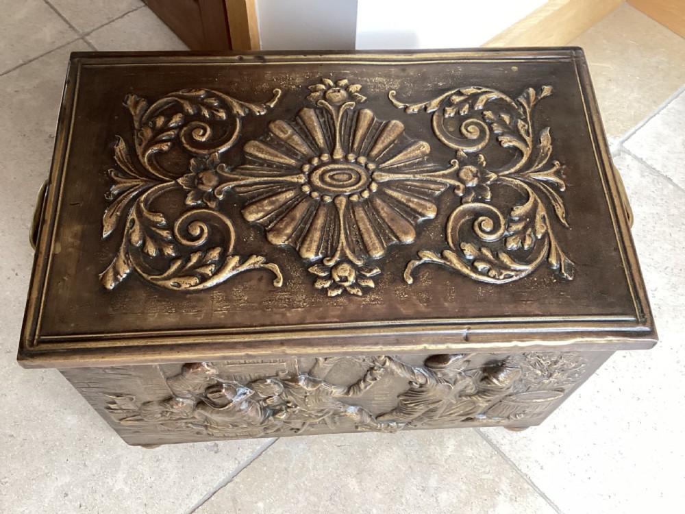 An antique brass-clad wooden fireside log box featuring embossed relief scenes of figures in a tavern on the front and foliate scrollwork on the lid. View VII