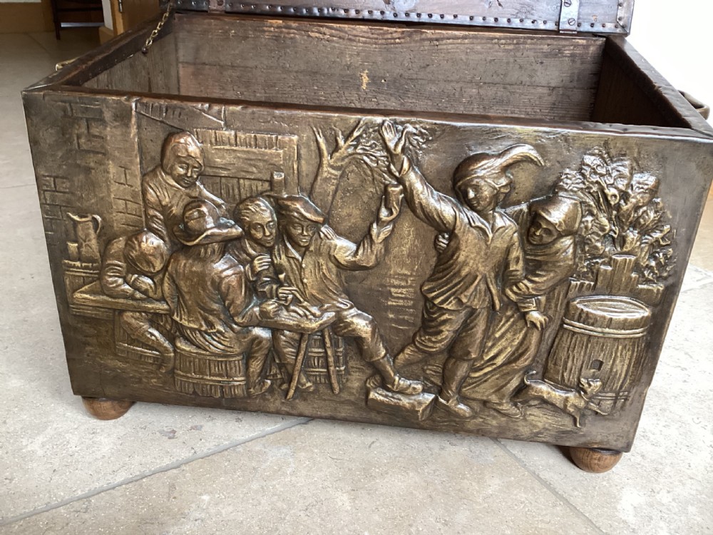 An antique brass-clad wooden fireside log box featuring embossed relief scenes of figures in a tavern on the front and foliate scrollwork on the lid. View XIV