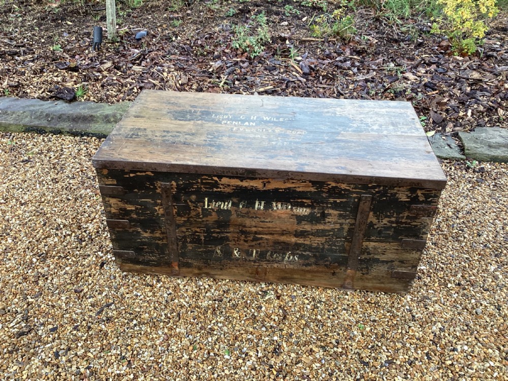 A rectangular antique dark wood military campaign chest featuring stencilled white lettering and iron strap reinforcements.