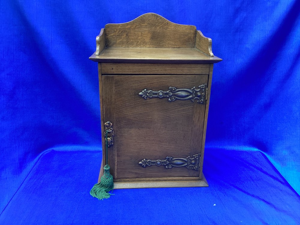 An Arts and Crafts medium brown oak key cabinet featuring an arched shaped gallery top and elaborate metal strap hinges on the single cupboard door.