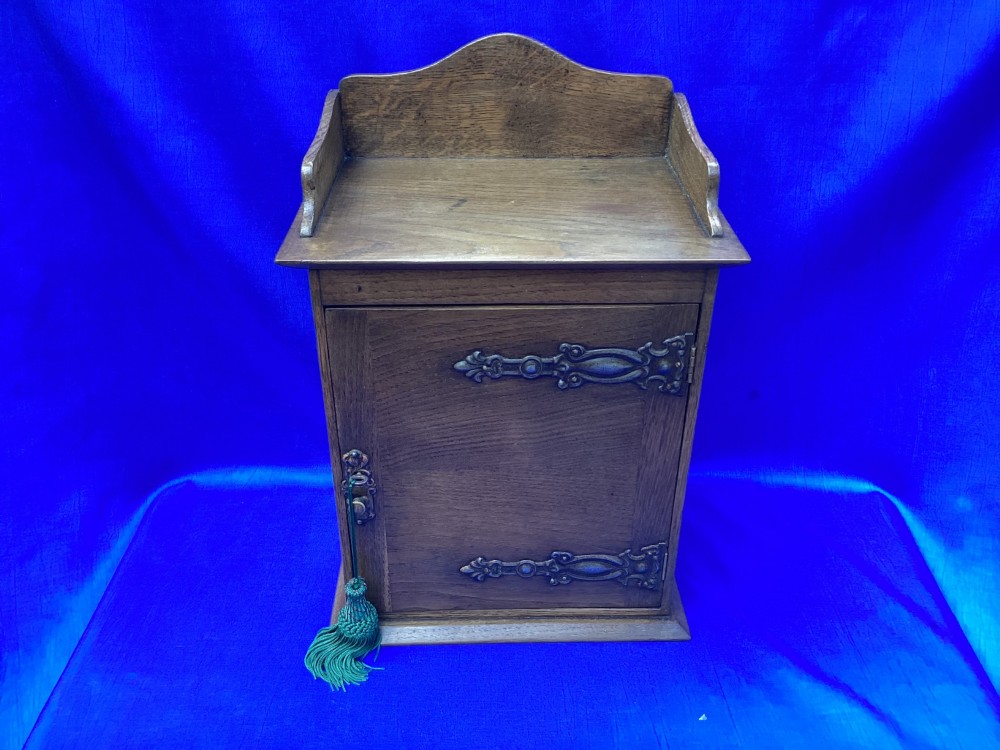 An Arts and Crafts medium brown oak key cabinet featuring an arched shaped gallery top and elaborate metal strap hinges on the single cupboard door. View II