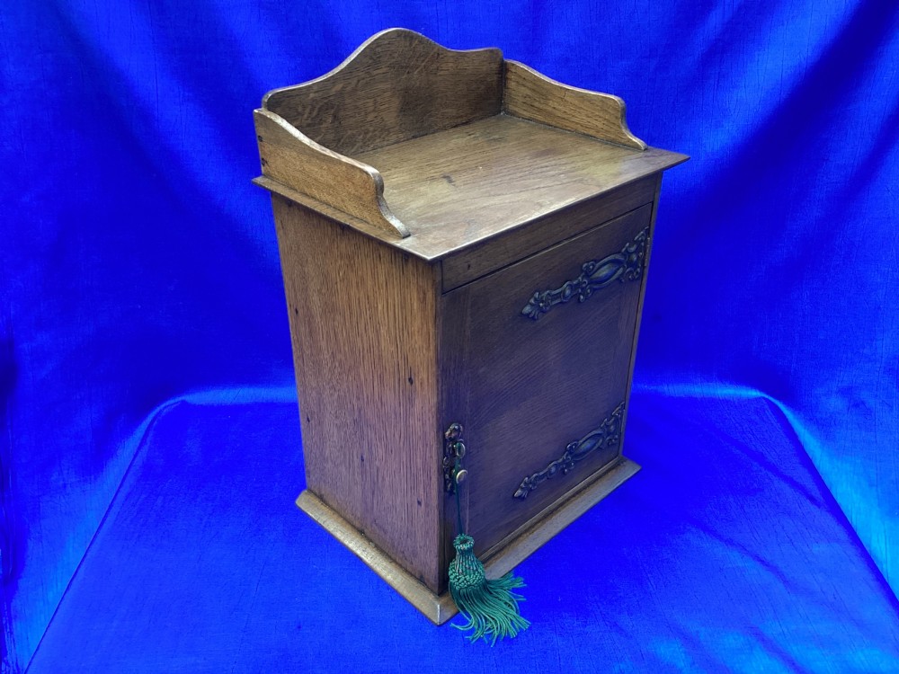 An Arts and Crafts medium brown oak key cabinet featuring an arched shaped gallery top and elaborate metal strap hinges on the single cupboard door. View IV