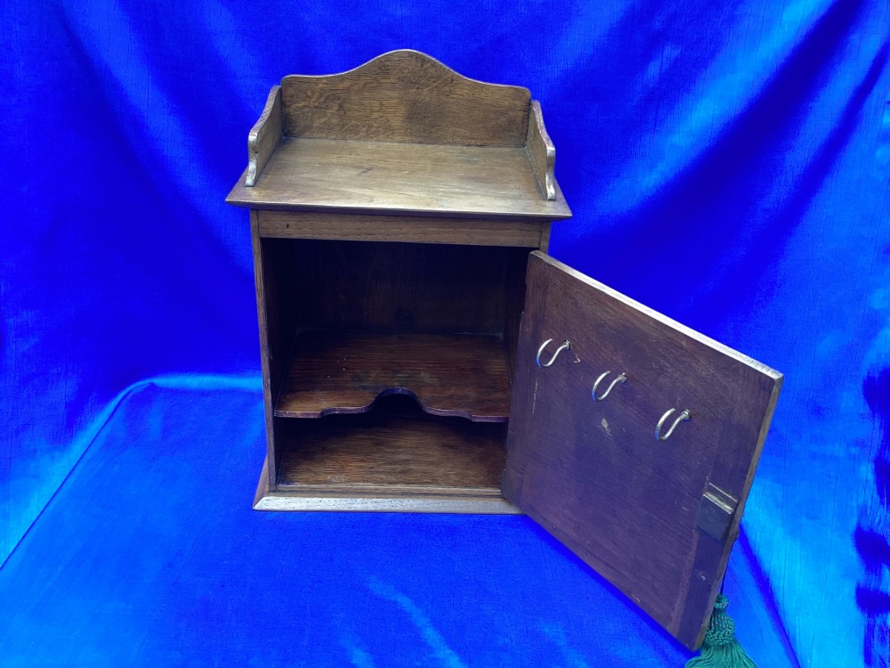 An Arts and Crafts medium brown oak key cabinet featuring an arched shaped gallery top and elaborate metal strap hinges on the single cupboard door. View V
