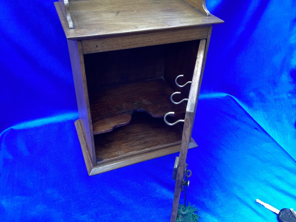 An Arts and Crafts medium brown oak key cabinet featuring an arched shaped gallery top and elaborate metal strap hinges on the single cupboard door. View VII