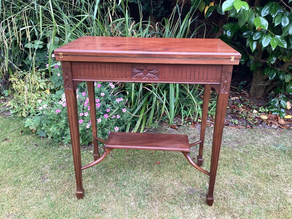An Edwardian mahogany fold-over games table featuring a carved floral frieze, fluted apron, slender tapered legs, and a lower tier shelf supported by swept stretchers.