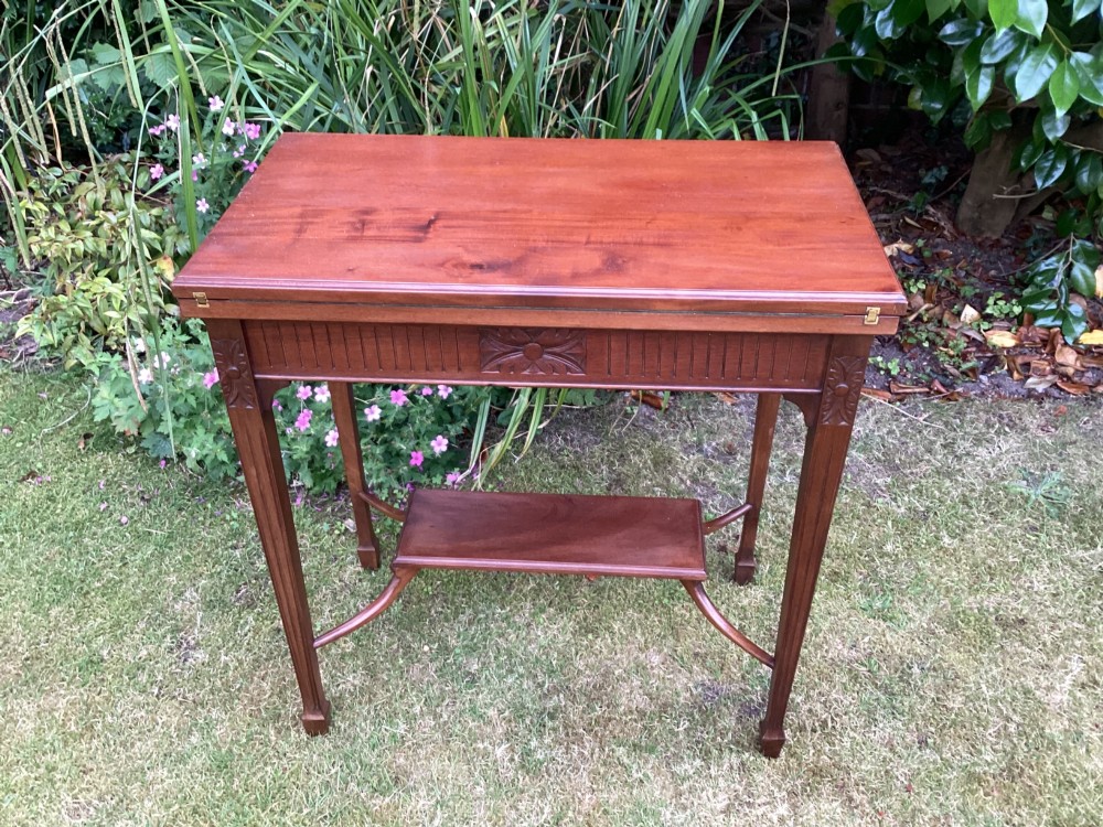 An Edwardian mahogany fold-over games table featuring a carved floral frieze, fluted apron, slender tapered legs, and a lower tier shelf supported by swept stretchers. View II
