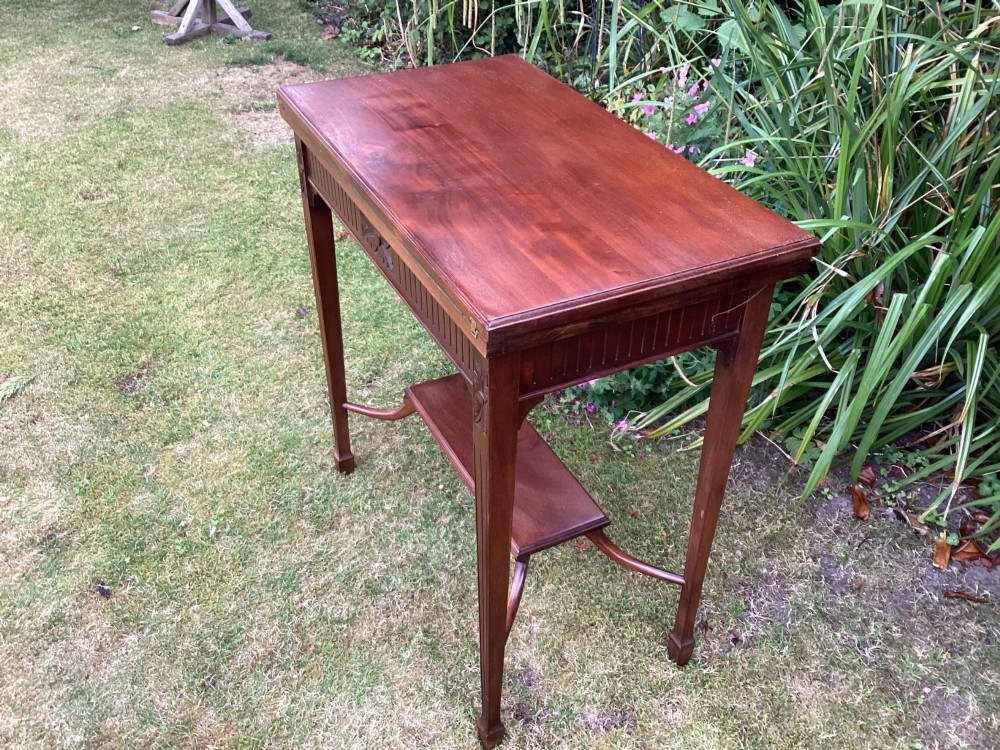 An Edwardian mahogany fold-over games table featuring a carved floral frieze, fluted apron, slender tapered legs, and a lower tier shelf supported by swept stretchers. View V