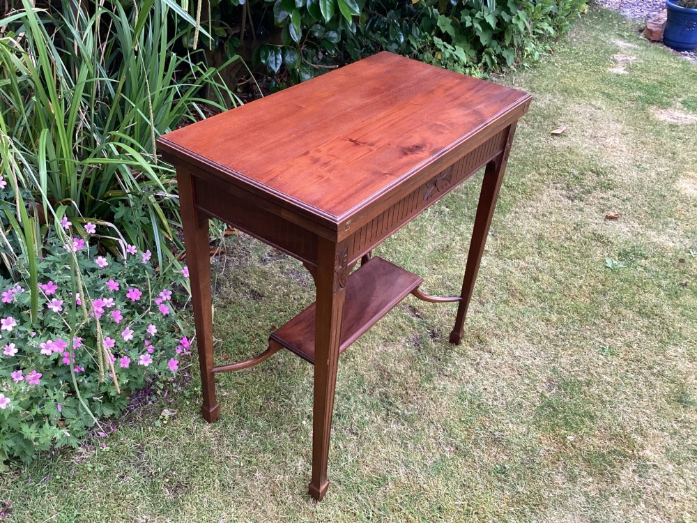 An Edwardian mahogany fold-over games table featuring a carved floral frieze, fluted apron, slender tapered legs, and a lower tier shelf supported by swept stretchers. View VI