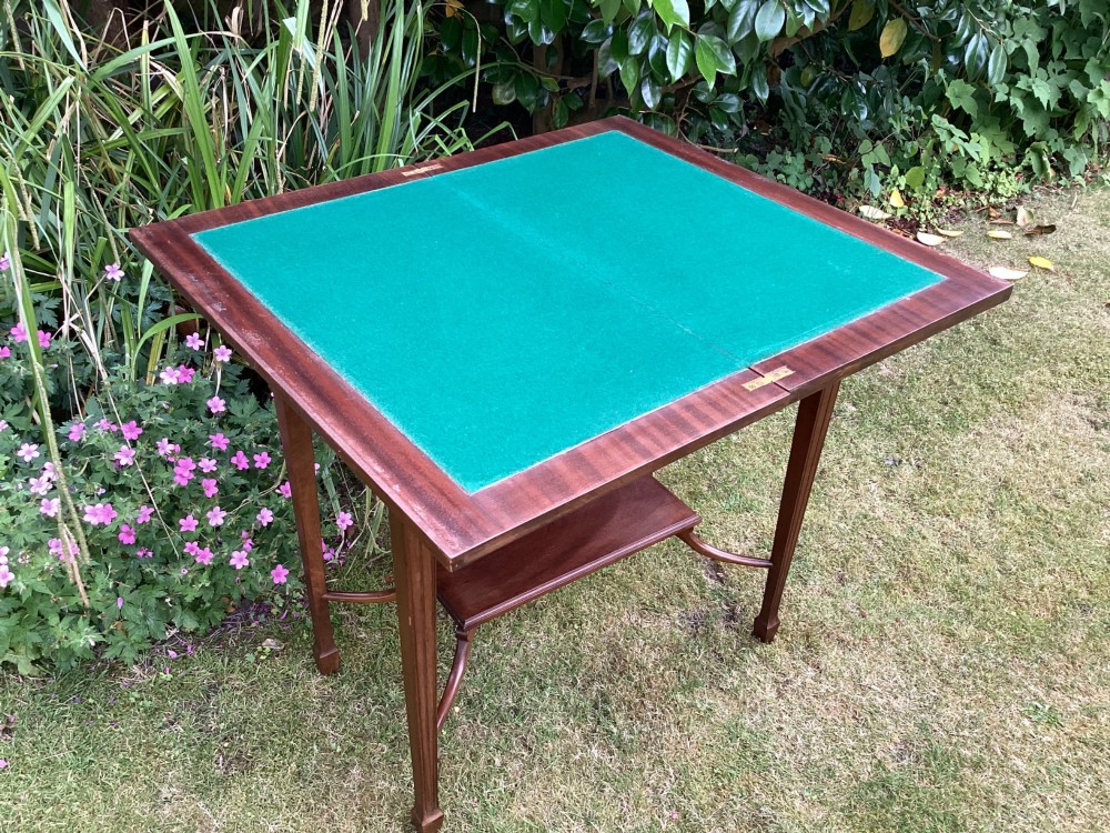 An Edwardian mahogany fold-over games table featuring a carved floral frieze, fluted apron, slender tapered legs, and a lower tier shelf supported by swept stretchers. View VII