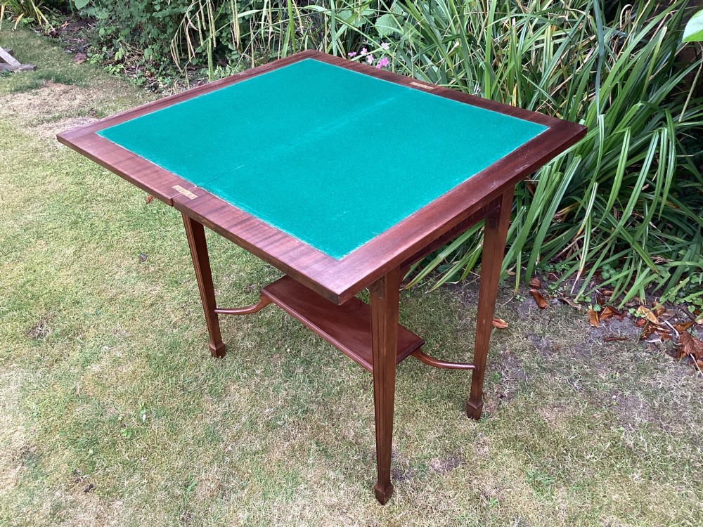 An Edwardian mahogany fold-over games table featuring a carved floral frieze, fluted apron, slender tapered legs, and a lower tier shelf supported by swept stretchers. View VIII