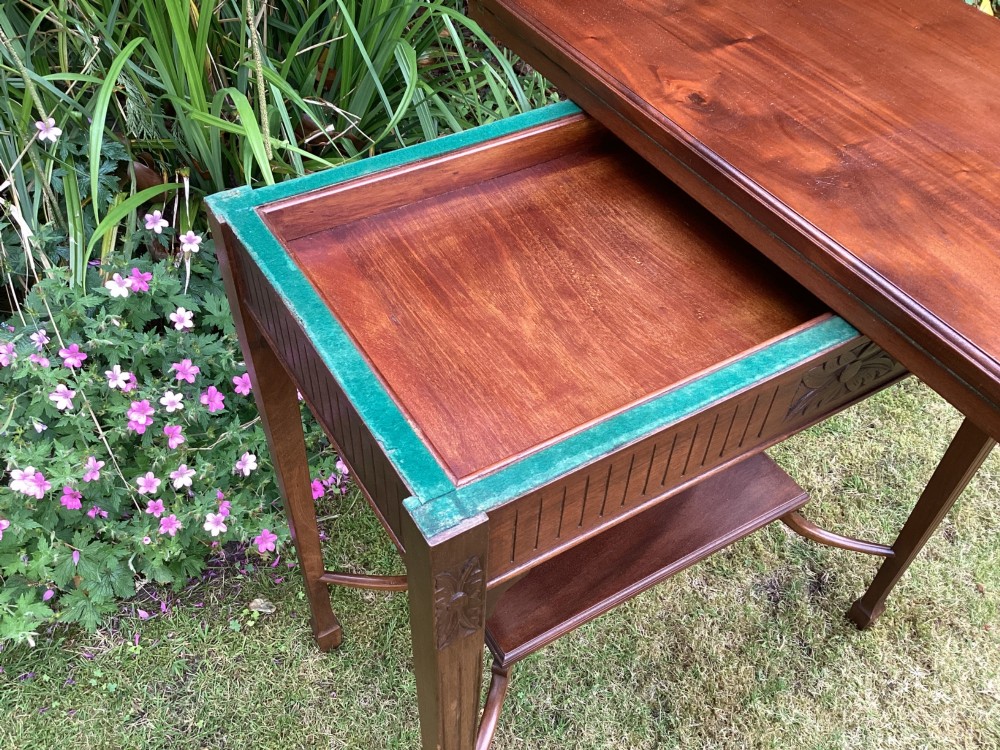 An Edwardian mahogany fold-over games table featuring a carved floral frieze, fluted apron, slender tapered legs, and a lower tier shelf supported by swept stretchers. View IX