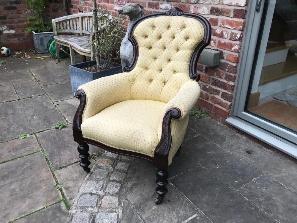 A Victorian mahogany parlour armchair featuring button-tufted pale yellow patterned upholstery, carved scroll arms, and turned front legs on castors. View V