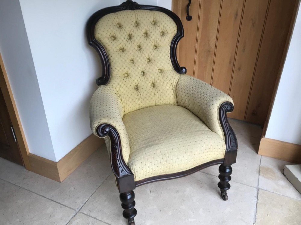 A Victorian mahogany parlour armchair featuring button-tufted pale yellow patterned upholstery, carved scroll arms, and turned front legs on castors. View VI