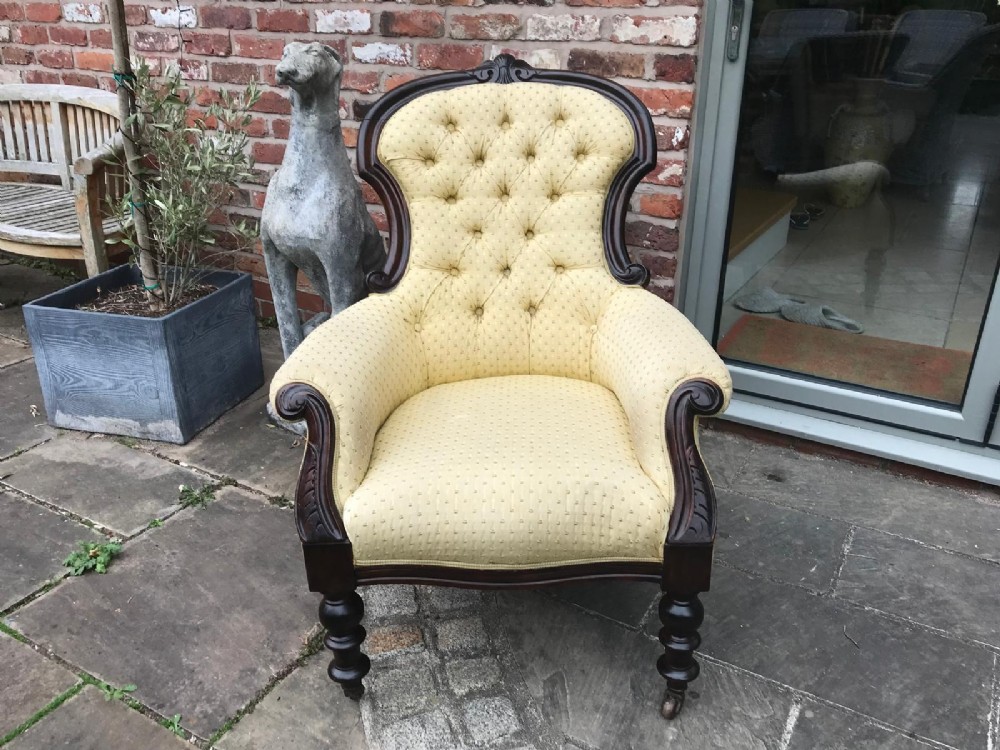 A Victorian mahogany parlour armchair featuring button-tufted pale yellow patterned upholstery, carved scroll arms, and turned front legs on castors. View VII