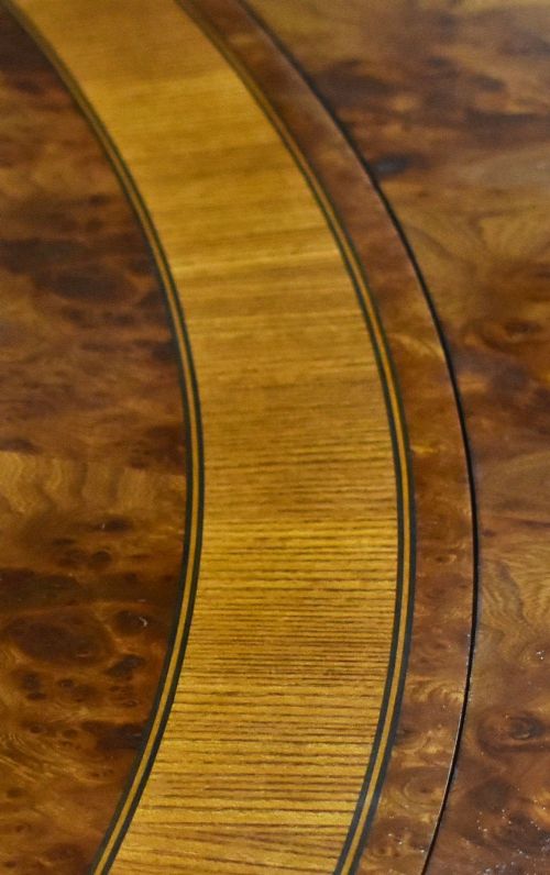 A large oval burr walnut extending dining table featuring decorative satinwood crossbanding and pedestal legs fitted with brass castors. View X