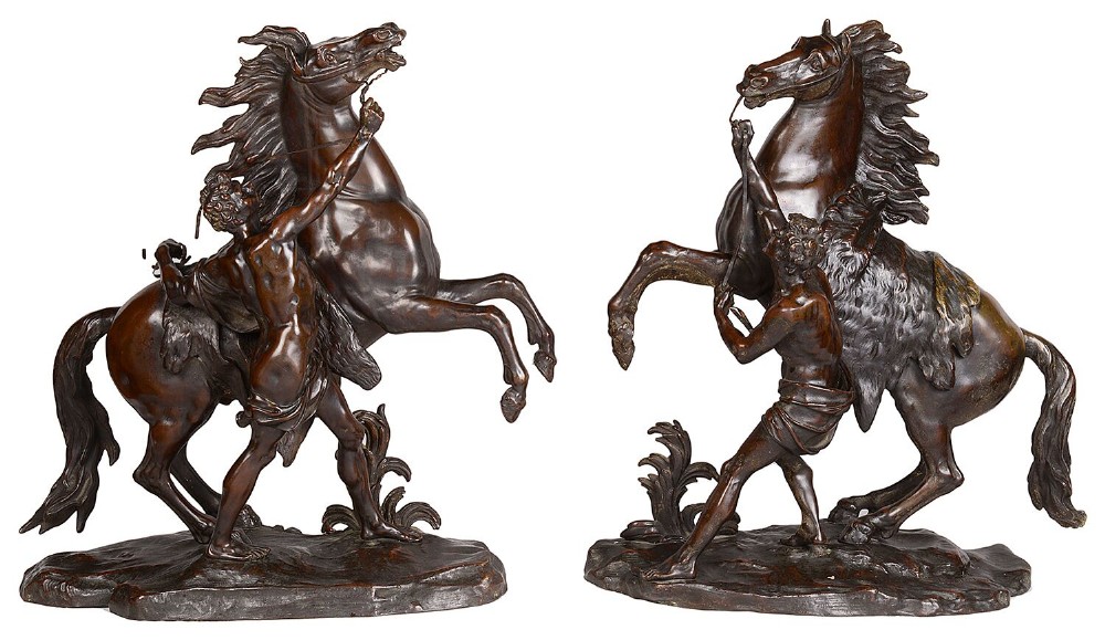 A pair of dark brown patinated bronze sculptures depicting rearing horses restrained by grooms on naturalistic bases.