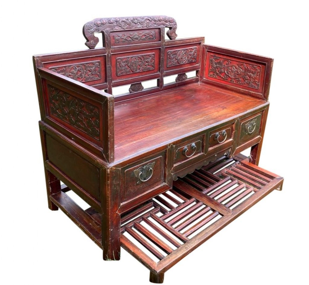 A red lacquered antique Chinese carved wooden sofa with carved back panels, storage drawers featuring brass drop handles, and an extended slat footrest.