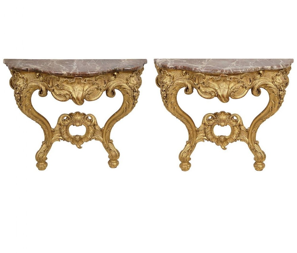 A pair of 19th-century French carved giltwood console tables featuring ornate Rococo scrollwork legs and reddish-brown marble tops.