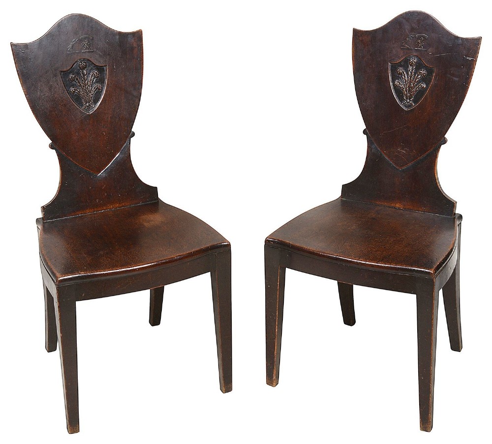 A pair of 18th-century mahogany hall chairs featuring carved shield backs with Prince of Wales feathers, solid wooden seats, and tapered square legs.