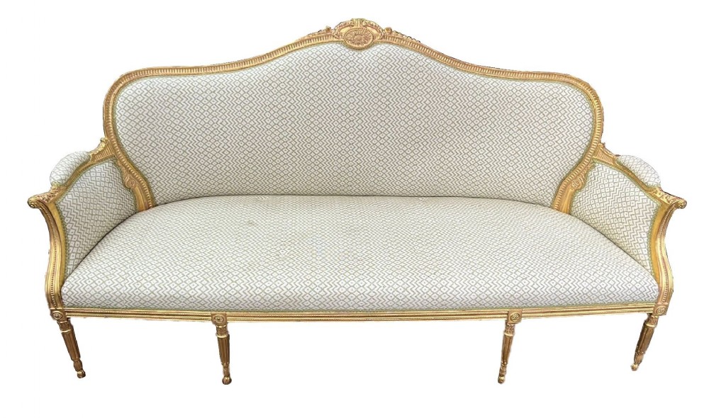 An antique Louis XVI style giltwood sofa featuring a carved gold leaf frame, fluted legs, and patterned cream upholstery.