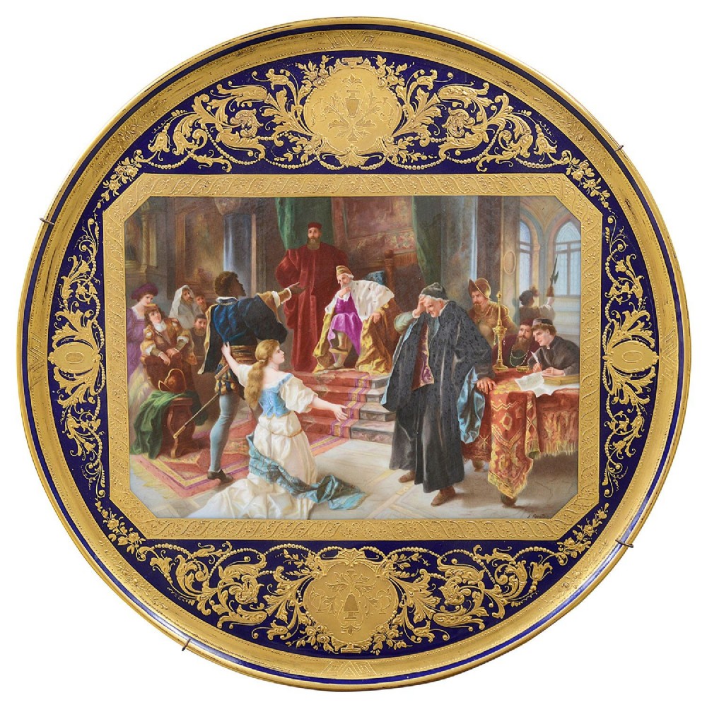 A large circular porcelain charger featuring a dark cobalt blue rim with intricate gilt scrollwork surrounding a central hand-painted scene depicting Othello.