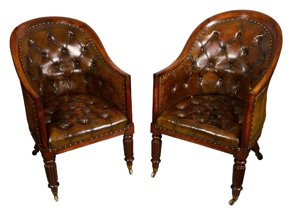 A pair of Regency period library armchairs featuring deep buttoned brown leather upholstery, curved tub backs, reeded front legs, and brass castors.