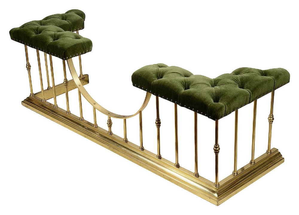 A Victorian brass club fender featuring vertical cylindrical spindles, a dipped front rail, and deep-buttoned green velvet corner seats with metal stud detailing.