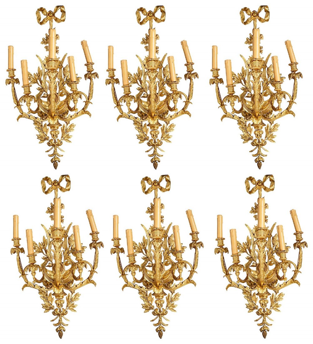 A set of six Louis XVI style gilded ormolu wall sconces featuring ribbon-bow finials and multi-arm candle holders adorned with leaf motifs.