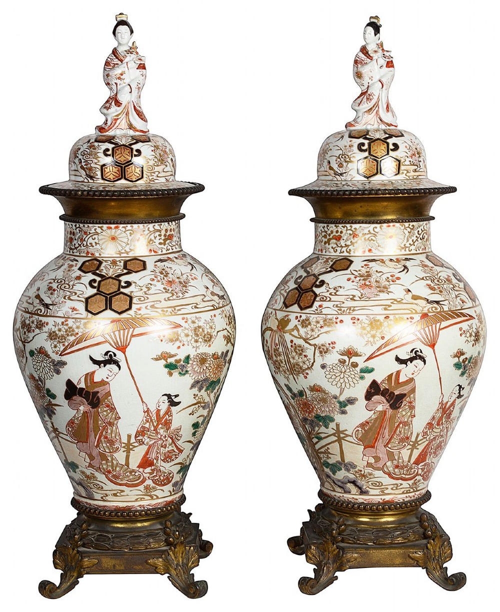Pair of Japanese Arita Imari porcelain lidded vases featuring painted figures under umbrellas, standing on bronze bases with standing figure finials.
