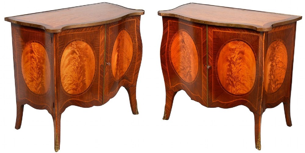 A pair of early 20th-century serpentine mahogany and satinwood commodes with oval central inlays and brass feet.