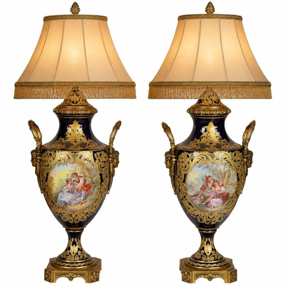 A pair of 19th-century French Sevres-style cobalt blue porcelain urn lamps featuring hand-painted romantic scenes, ornate gilt bronze mounts, and fringed fabric shades.