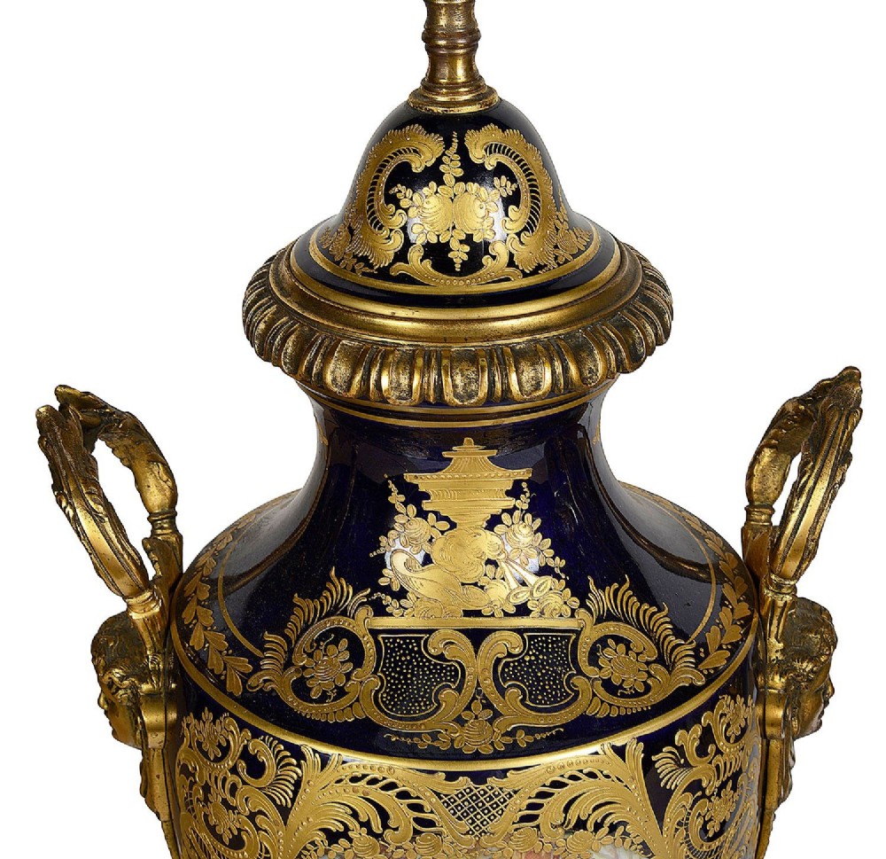 A pair of 19th-century French Sevres-style cobalt blue porcelain urn lamps featuring hand-painted romantic scenes, ornate gilt bronze mounts, and fringed fabric shades. View VI