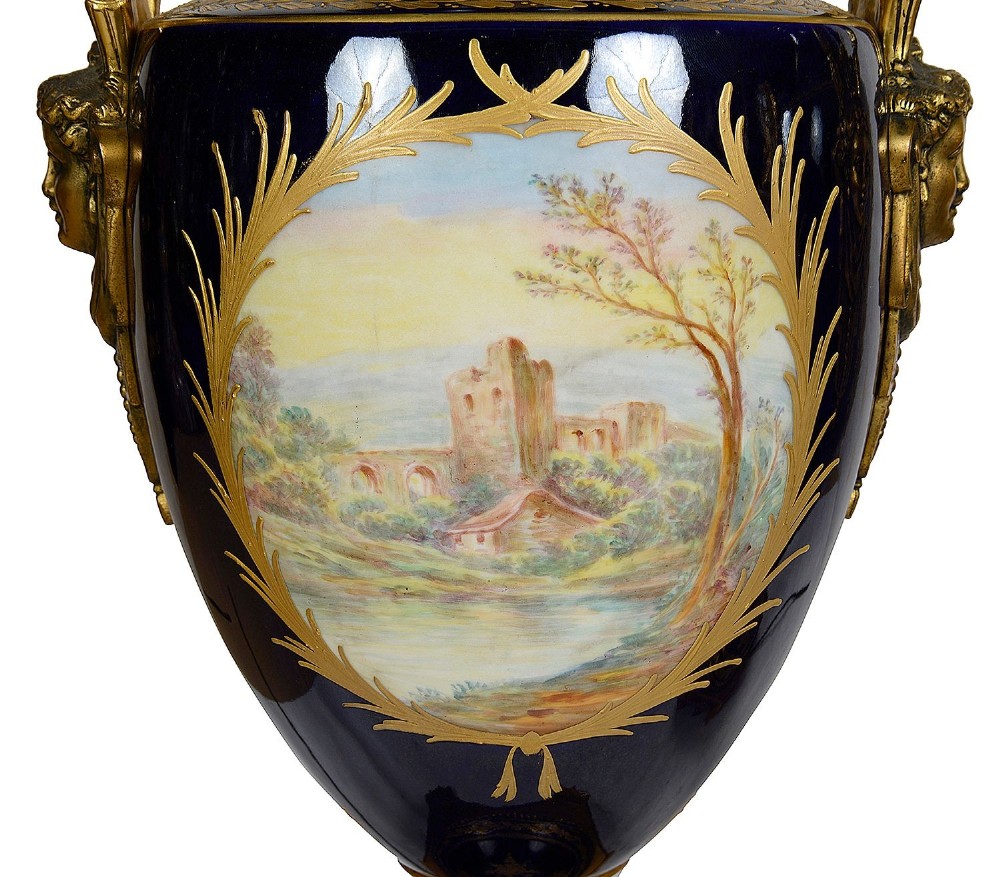 A pair of 19th-century French Sevres-style cobalt blue porcelain urn lamps featuring hand-painted romantic scenes, ornate gilt bronze mounts, and fringed fabric shades. View X