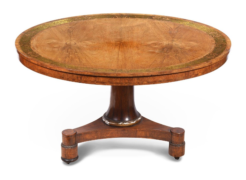 A circular Regency period timber centre table featuring a floral brass inlaid border, a central tapered pedestal stem, and a tripartite base resting on cylindrical feet with castors. View II