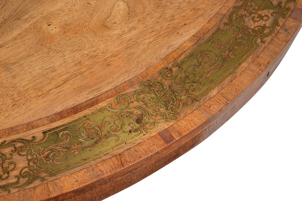A circular Regency period timber centre table featuring a floral brass inlaid border, a central tapered pedestal stem, and a tripartite base resting on cylindrical feet with castors. View IV