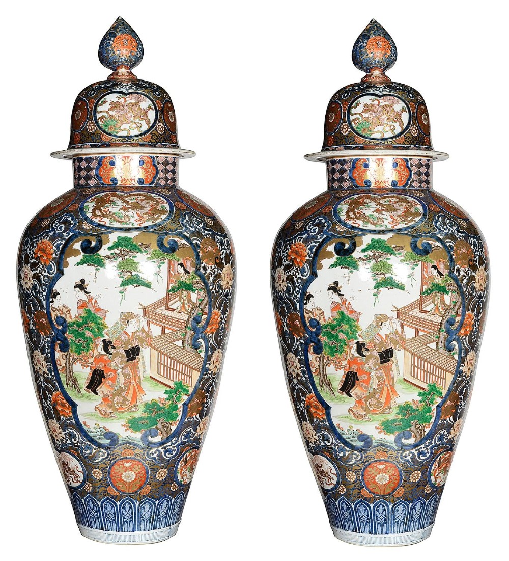 A pair of large 19th-century Japanese Imari porcelain lidded vases featuring painted reserves depicting figures in garden settings, surrounded by cobalt blue, orange, green, and gold patterns.