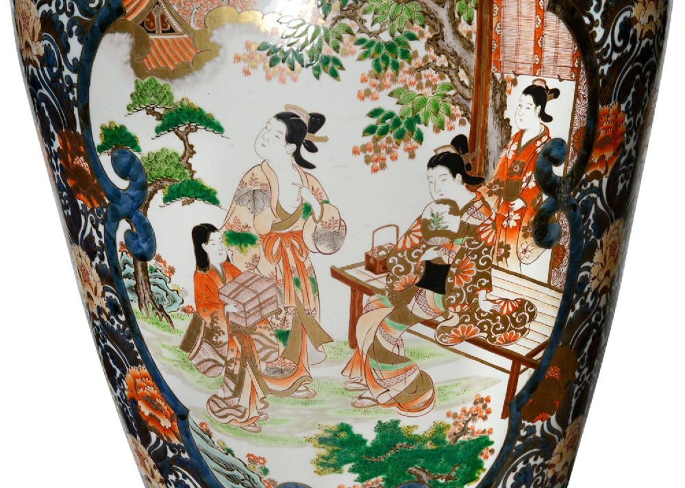 A pair of large 19th-century Japanese Imari porcelain lidded vases featuring painted reserves depicting figures in garden settings, surrounded by cobalt blue, orange, green, and gold patterns. View X