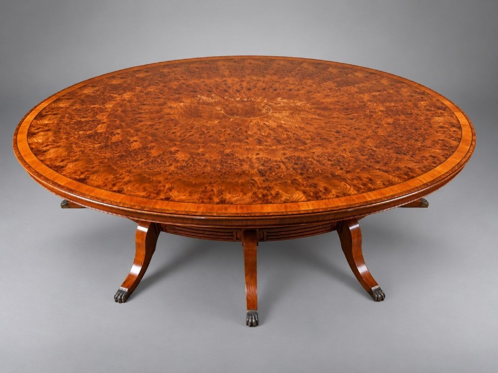A large oval burr walnut extending dining table featuring decorative satinwood crossbanding and pedestal legs fitted with brass castors. View II