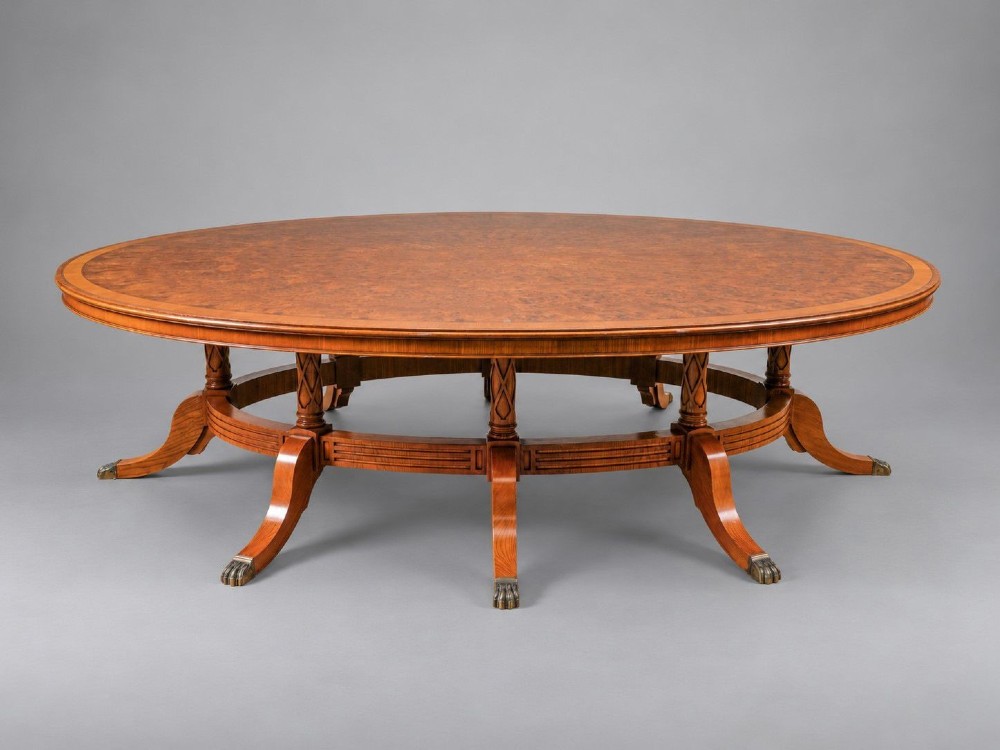 A large oval burr walnut extending dining table featuring decorative satinwood crossbanding and pedestal legs fitted with brass castors. View III
