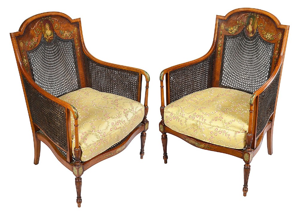 A pair of late Victorian Sheraton Revival satinwood bergere armchairs featuring hand-painted floral motifs, portrait medallions, caned backs, and yellow fabric seats.