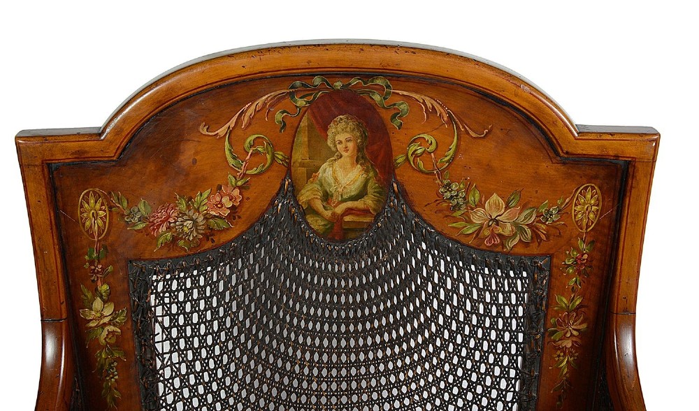 A pair of late Victorian Sheraton Revival satinwood bergere armchairs featuring hand-painted floral motifs, portrait medallions, caned backs, and yellow fabric seats. View V
