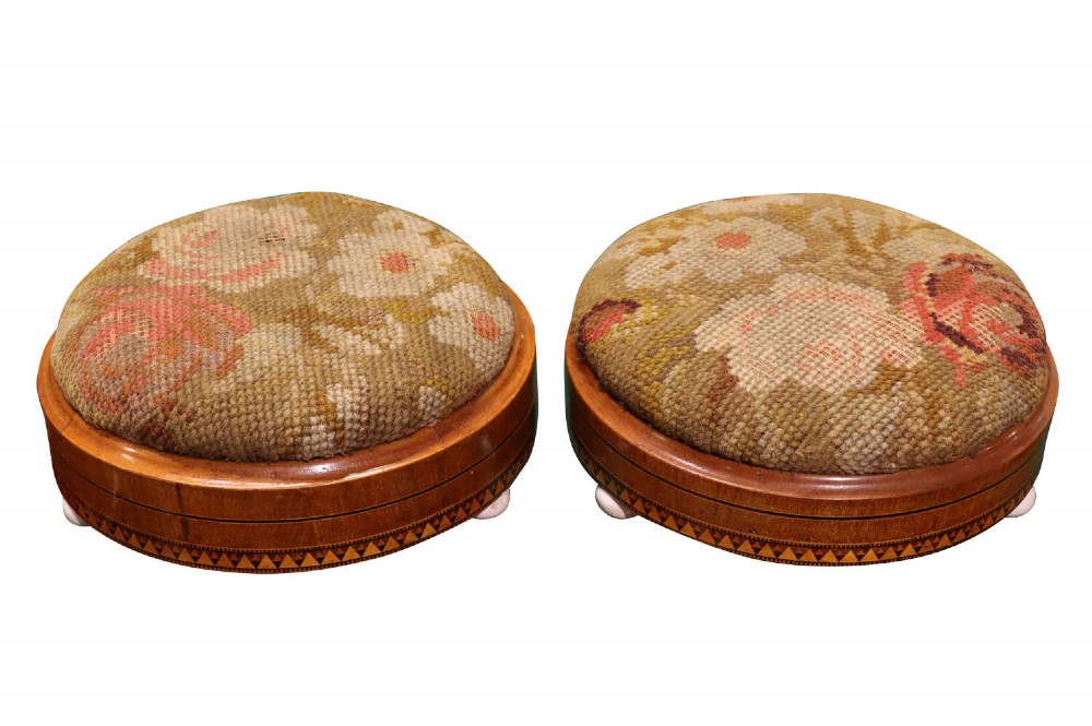 A pair of circular Victorian walnut footstools featuring floral needlepoint cushions and decorative geometric inlaid wood borders, elevated on small ceramic bun feet.