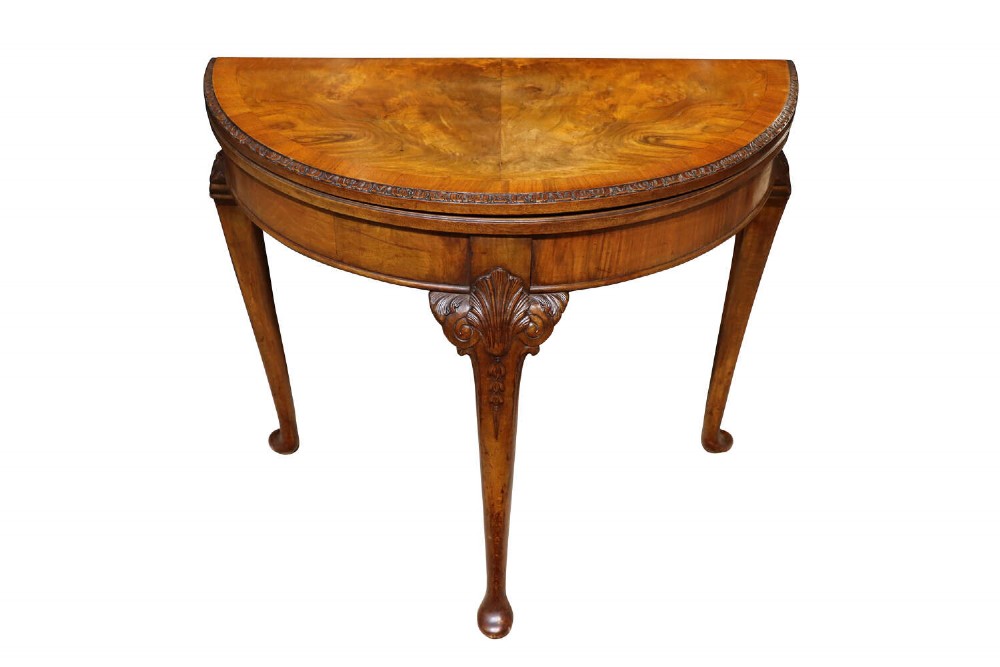 A 1920s Queen Anne style demi-lune card table crafted from burr walnut featuring cabriole legs with carved shell details and pad feet.